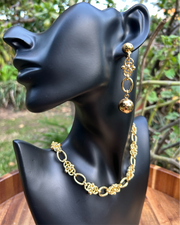 A pair of gold-colored hoop earrings displayed on a black mannequin head, set against a backdrop of greenery.