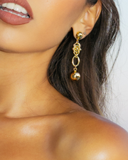 Close-up of a person wearing gold DIPHDA Eclipse Earrings  with a blurred background - Half ball stud with a handmade unique interwoven large chain with a hollow golden ball hanging from end- looks expensive yet edgy, classic yet modern. 